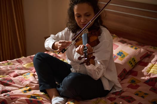 A woman in a casual white shirt plays the violin while sitting on a cozy bed, expressing concentration and passion for music.