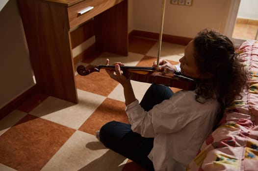 A woman practicing the violin while sitting on the floor in a comfortable room, focused and serene. The warm lighting adds to the peaceful atmosphere of the scene.