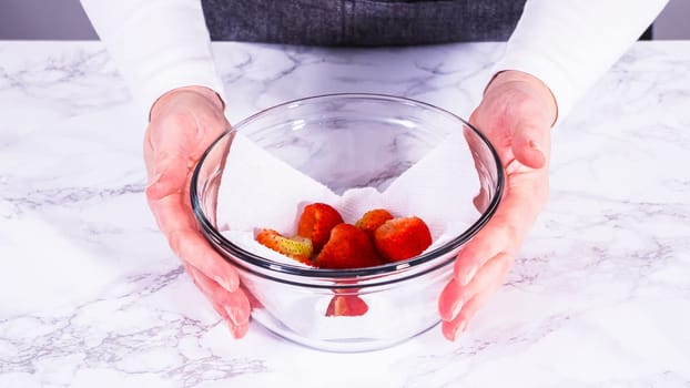Strawberries, freshly washed and dried, are neatly stored in a glass bowl lined with a paper towel and securely covered with plastic wrap to maintain freshness.
