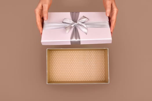 Female hands opening an empty gift box adorned with a silver ribbon bow set against a warm brown Mocha Mousse color of 2025 year background capturing the essence of surprise and celebration from above