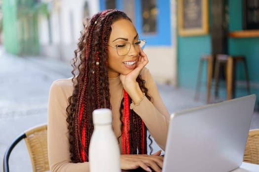 A young woman with braided hair smiles while using her laptop at an outdoor caf, showcasing a relaxed but productive atmosphere