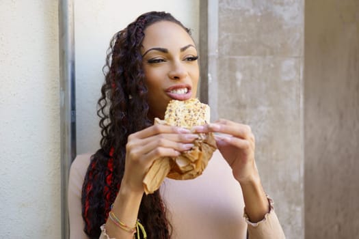 A cheerful young woman with curly hair beams with delight as she takes a satisfying bite of a delicious snack that is wrapped in paper, cherishing her wonderful moment spent outdoors in nature