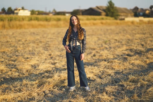 A confident young woman stands in a sunlit field, dressed in trendy flared jeans and a vibrant top, surrounded by a picturesque landscape that enhances her cheerful spirit and youthful energy