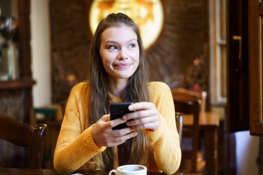 A cheerful young woman casually holding a smartphone while seated at a caf table, enjoying a moment of relaxation