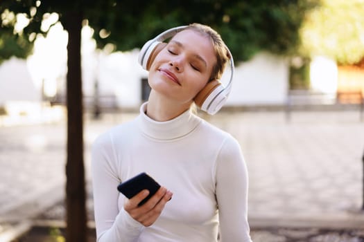 A young woman dressed in a cozy white sweater is blissfully listening to her favorite music through headphones while holding a smartphone outdoors, enjoying the beautiful surroundings of nature