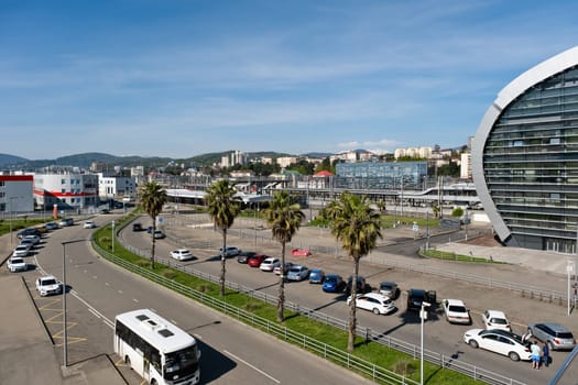 City view with palm trees and modern train station building next to busy parking lot and roadway in sunny weather - Sochi, Russia April 16, 2024.