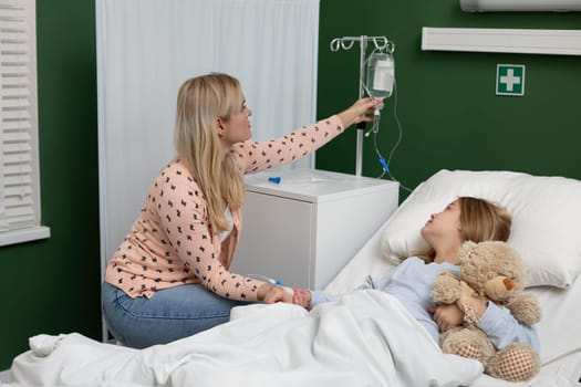 In a hospital, a caring mother supports her child with an IV, who holds a beloved teddy bear for comfort