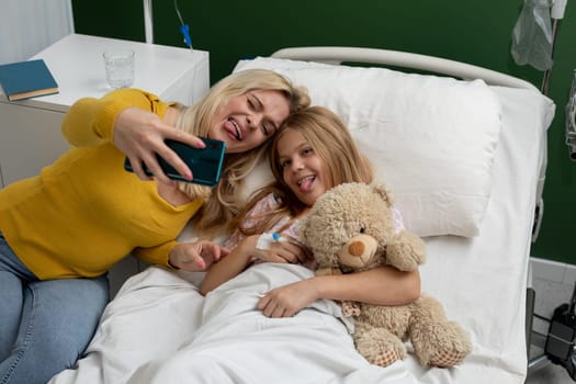 A cheerful mother and her adorable daughter are capturing a silly and joyful selfie while in a hospital bed