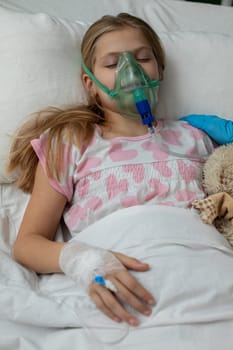 A young girl is resting peacefully in a hospital bed while using an oxygen mask and hugging her favorite teddy bear