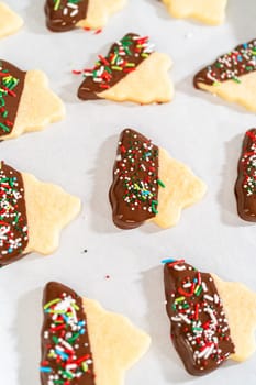 Buttery cookies half-dipped in chocolate, sprinkled with festive red, green, and white decorations, laid on white parchment to set.