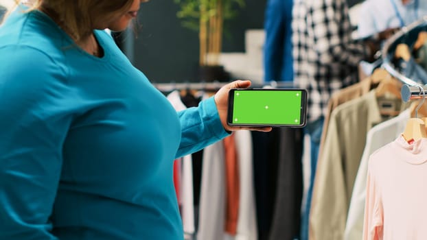 African american woman analyzing phone with greenscreen template, looking at isolated display in modern boutique. Stylish customer shopping for trendy outfit in clothing store. Fashion concept