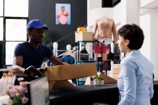 African american courier taking cardbord boxes from store worker, discussing shipping details in shopping mall. Employee preparing packages for delivery, working in modern boutique