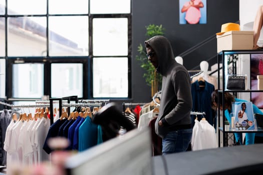 African american man wearing hood and glasses trying to steal fashionable clothes from modern boutique. Robber looking around to see if someone watching him while stealing merchandise