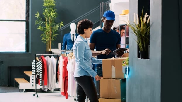 Manager helping courier with packages, carrying cardboard boxes in modern boutique. African american deliveryman explaining shipping details to worker in clothing store. Fashion concept