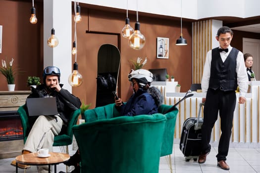 In winter ski resort lounge couple looking at screen of their digital devices while surrounded by snowboarding gear. Guests seated comfortably with laptop and smartphone while bellboy takes suitcase.