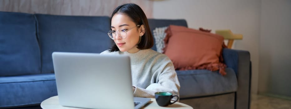 Self employed young korean woman working on remote, typing on laptop, studying at home in living room.