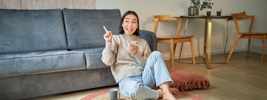 Portrait of stylish korean woman sits on floor with smartphone, using mobile phone, smiling pleased, concept of staying at home and relaxation.