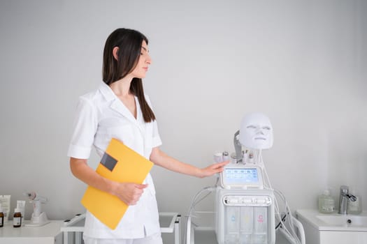 Woman cosmetologist standing in medical office. Female dermatologist holding yellow folder with patient medical history standing near modern equipment. Dermatology specialist in uniform at hospital