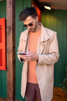 Fashionable man in coat chatting outside. Elegant dark haired male reading text message on his phone. Online communication: handsome man texting with cell phone during Christmas holidays