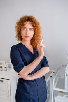 Female cosmetologist at medical office. Redhead woman dermatologist wearing scrubs holds an ampule at hospital. Skin care practitioner in navy blue medical uniform stands in aesthetic clinic office