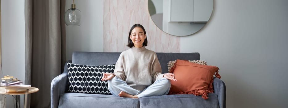 Relaxation and patience. Smiling young asian woman in cozy room, sitting on sofa and meditating, doing yoga mindfulness training.