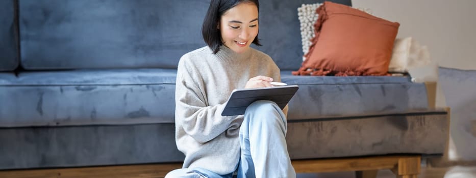 Vertical shot of cute asian girl in cozy home, drawing on her digital tablet with pen, designer working on project.