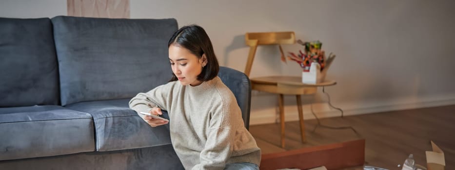 Vertical shot of young woman in cozy home working on laptop, using smartphone and drinking coffee, sitting on floor near sofa.