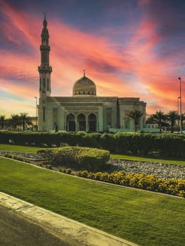 A photo of a white mosque with a dome and a minaret at sunset. The mosque is surrounded by a green garden with yellow flowers. The image has a low angle and an orange and pink sky. High quality photo