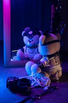 the toy bear in leather harness and accessory for BDSM games on a dark background in neon light near the mirror