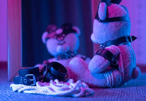 the toy bear in leather harness and accessory for BDSM games on a dark background in neon light near the mirror