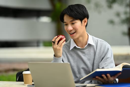 Happy young asian man eating apple and using laptop at campus. Technology, education and lifestyle concept.
