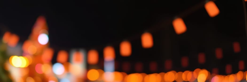 Vibrant orange lights hang over a lively night market, creating a festive atmosphere. The background features blurred city buildings and bustling activity, enhancing the celebrations.