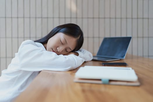 A young woman rests her head on her arms at a desk, feeling overwhelmed. The laptop beside her reflects a modern workspace, highlighting the need for rest and balance in daily life.