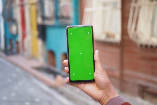 A hand grips a smartphone with a bright green screen in a lively, colorful street scene