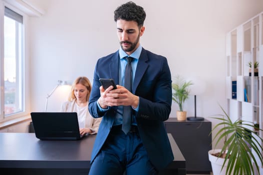 Businessman Young businessman in a blue suit checking his smartphone while leaning on a desk in a modern office. A female colleague works on a laptop in the background, creating a professional scene.