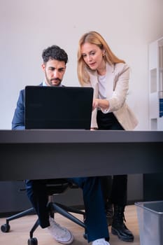 ProfeA businesswoman in a formal suit leans over to explain details on a smartphone to a seated female colleague working on a laptop. Vertical