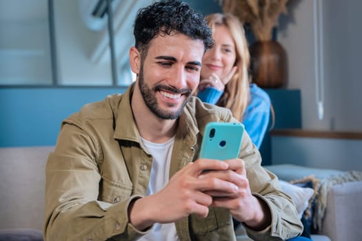 Smiling man using smaA cheerful man holds a smartphone while sitting in a cozy home environment. A smiling woman in the background watches him with interest.