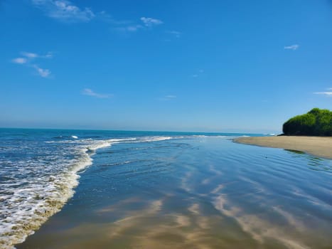 A tranquil beach with gentle waves and clear blue sky, featuring sandy shores and a distant greenery patch.