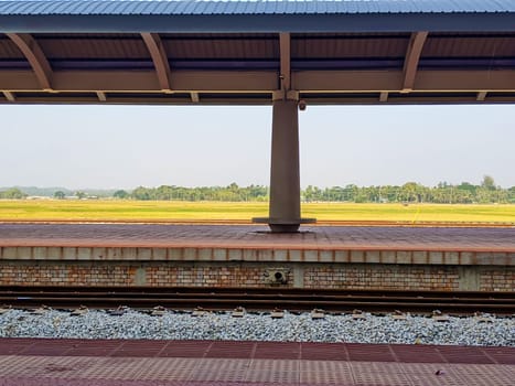 The image shows an empty train platform with a covered roof, surrounded by an open field with sparse trees in the distance.
