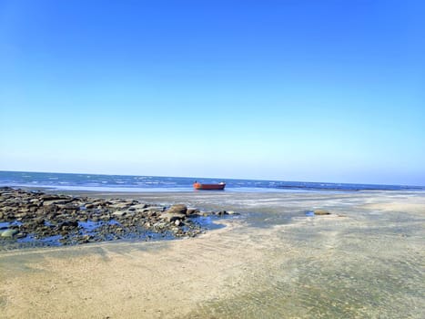 A red boat is stranded on a shallow, rocky shoreline with clear water and a vast, calm sea under a blue sky.