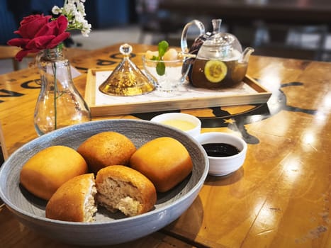 A cozy cafe scene with a bowl of freshly baked buns on a wooden table. There's also a teapot with a glass cup of tea, a vase with flowers, and a tray with a bowl of fruit.