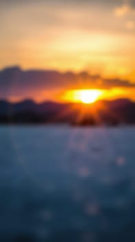 A picturesque sunset casts warm golden and orange shades across the sky, silhouetting distant mountains. The tranquil sea reflects the vibrant colors, blending with a soft blur.