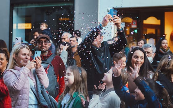 The image shows a group of people gathered in what appears to be a public outdoor setting. They are looking towards the same direction, some with smiles on their faces, indicating they are watching something that is likely of interest or entertainment. Several people are using their smartphones to take pictures or record the event. There is also confetti in the air, suggesting the occasion might be a celebratory event, like a festival, concert, parade, or a party. The atmosphere seems lively and joyful.