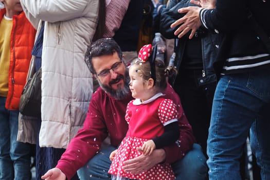 The image depicts a man and a young girl. The man is seated and has glasses and facial hair. He is wearing a red shirt and blue jeans. He's looking at the young girl with a gentle expression, possibly a look of care or affection. The girl, who is standing and appears to be having a conversation with the man, is wearing a red dress with white polka dots, which is complemented by a headband with a bow that matches her dress. They seem to be enjoying a moment together amidst a background with other people, suggesting they could be at a public event or gathering. The image captures a candid, personal interaction between the two.