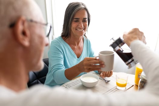 Happy mature couple having healthy breakfast together at home dining room. Man serving coffee to wife. Lifestyle concept.