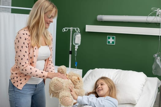A mother comforts her sick child in a hospital room by sharing a treasured teddy bear for emotional support