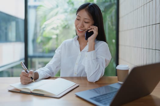 A cheerful young woman engages in a phone conversation while jotting down important notes, surrounded by a modern workspace, representing productivity and creativity.
