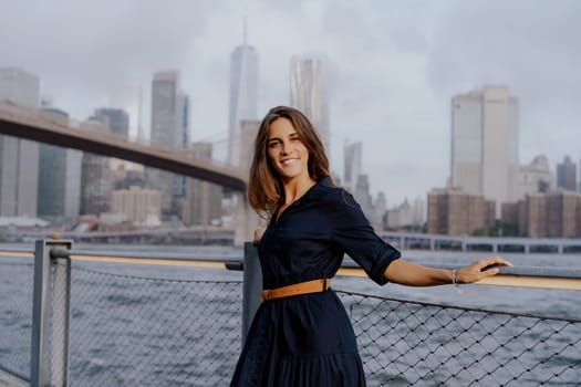 A stylish and fashionable woman confidently poses by the beautiful waterfront with an incredible city skyline view. Brooklyn bridge in New York.