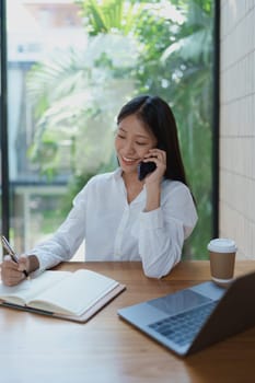 A young woman smiles while talking on the phone, jotting down notes in her notebook. A sleek laptop and coffee cup rest on the wooden table, creating an inviting workspace with greenery in view.