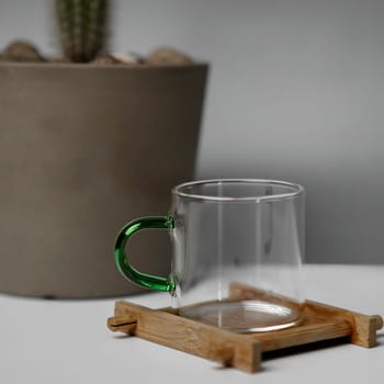 An elegant, minimalist glass mug is beautifully placed on a wooden coaster next to a green potted cactus plant